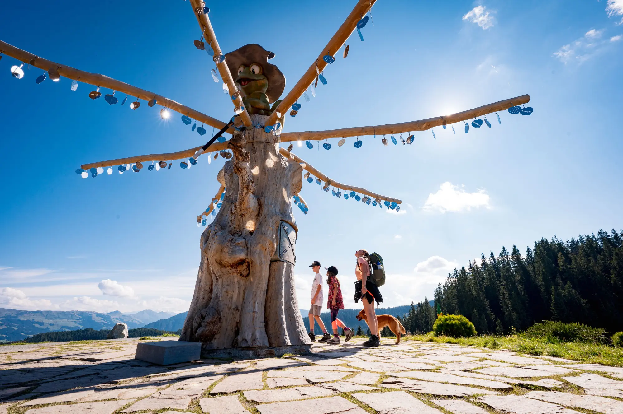Eine Gruppe von Personen mit einem Hund betrachtet eine künstlerische Baumskulptur mit einer Froschfigur unter blauem Himmel in Ellmi's Zauberwelt in Tirol.