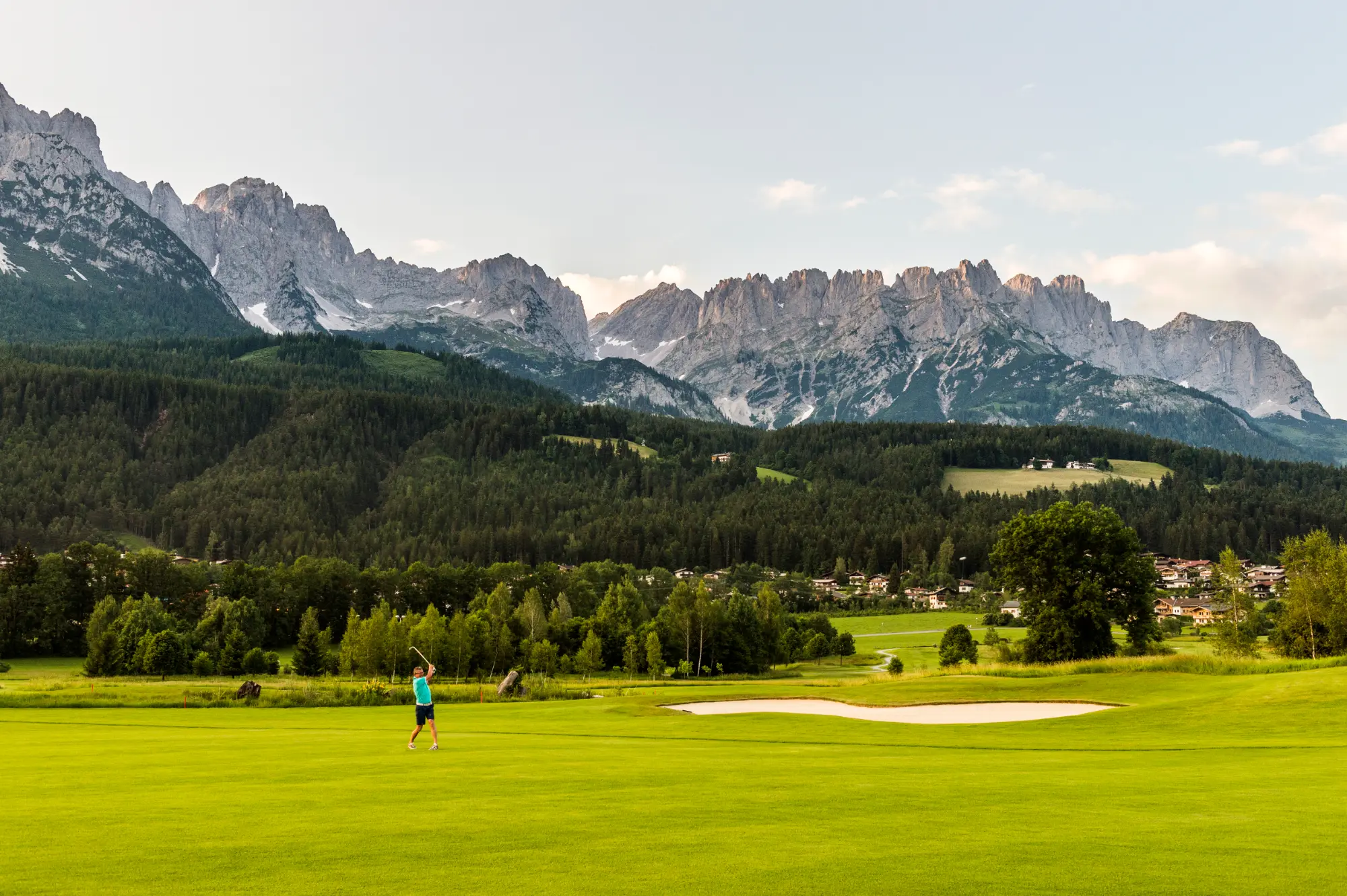 Ein Golfer beim dynamischen Abschlag auf dem intensiv grünen Rasen in Ellmau vor den mächtigen, grauen Felswänden des Kaisergebirges in Tirol.