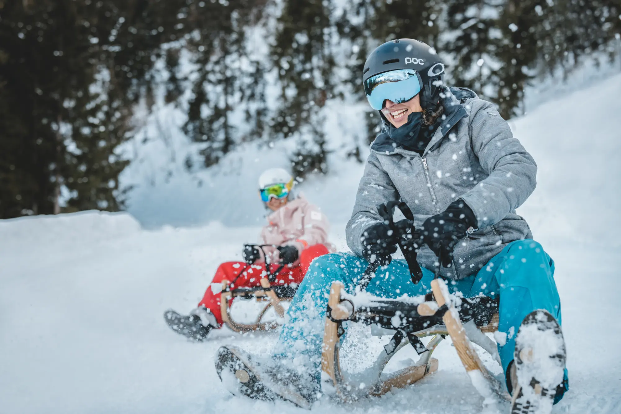 Eine lachende Frau mit POC-Helm und Skibrille beim rasanten Rodeln auf der winterlichen Naturrodelbahn am Astberg, während frisch aufgewirbelter Schnee spritzt.