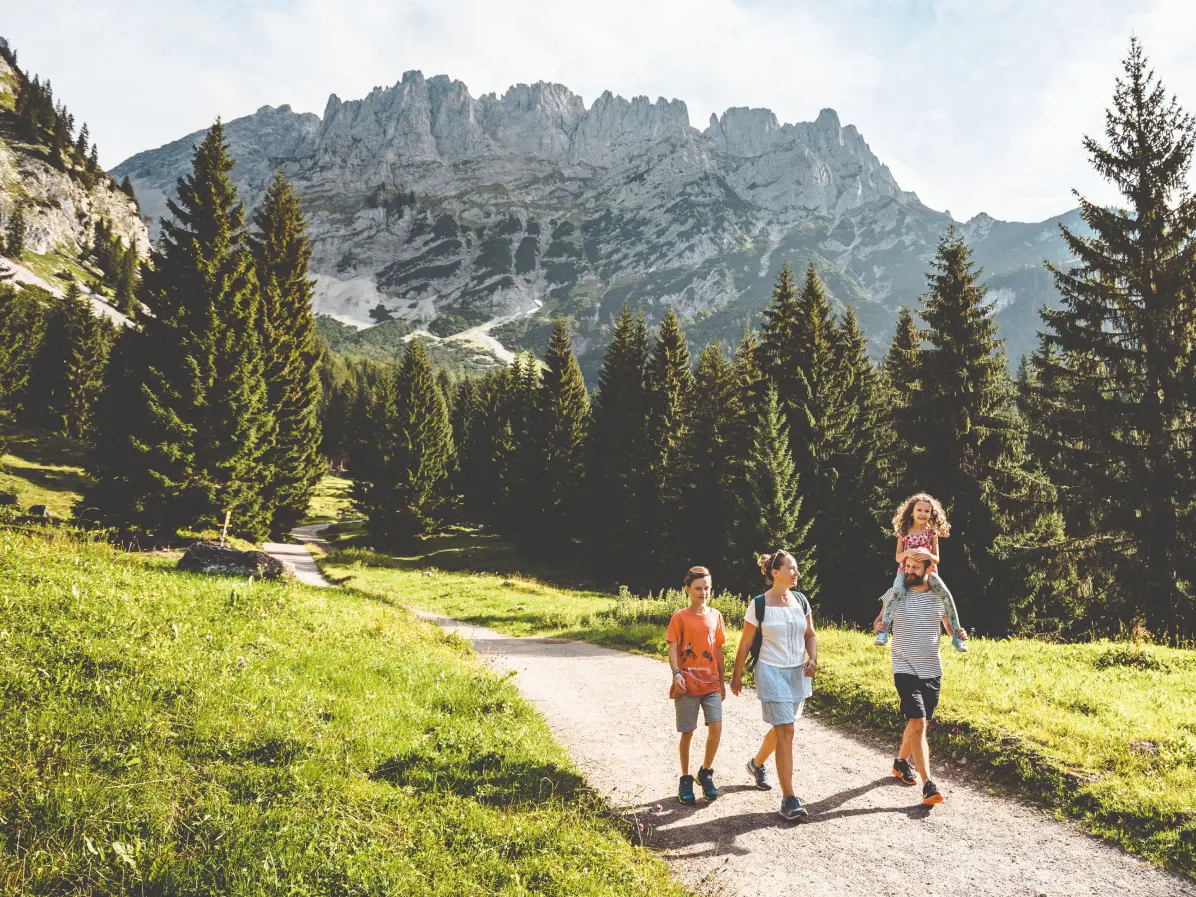 Eine vierköpfige Familie wandert an einem sonnigen Sommertag auf einem Schotterweg in der Region Wilder Kaiser in Tirol. Der Vater trägt die kleine Tochter auf den Schultern, im Hintergrund ragen dichte Nadelwälder und die schroffen Felswände des Kaisergebirges empor.