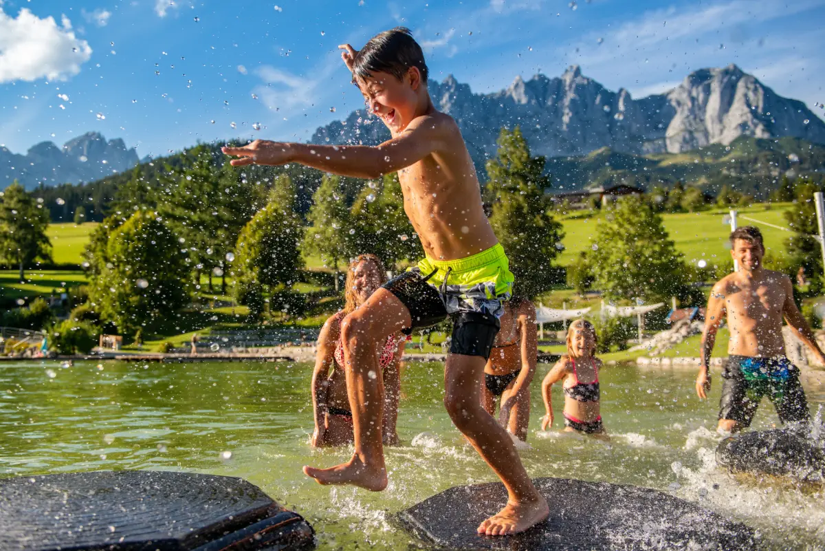 Kinder springen ins Wasser am Badesee Going, dahinter das Kaisergebirge